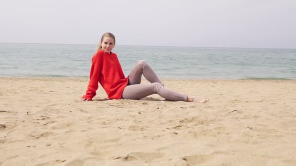 Menina loira está sentada na areia junto ao mar e posando para uma sessão de fotos — Vídeo de Stock