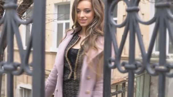 Дівчина в пальто позує за парканом на вулиці — стокове відео