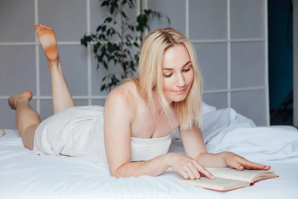 Hermosa mujer rubia lee un libro en el dormitorio en la cama —  Fotos de Stock