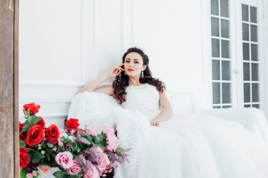 Gelinlik ve taç giymiş gelin beyaz salonda oturuyor.