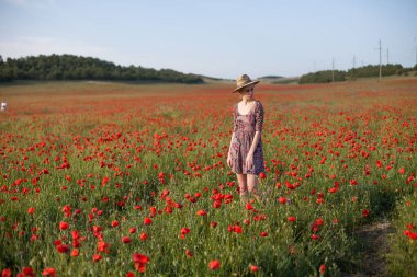 Kırmızı çiçeklerle dolu bir tarlada şapkalı şık, sarışın bir kadın.
