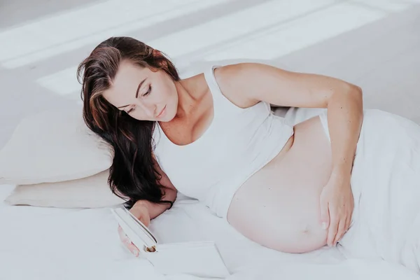 En gravid kvinna ligger på en säng och läsa en bok — Stockfoto