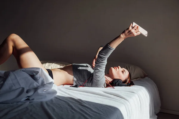 Een meisje neemt foto op een smartphone op het bed met een koptelefoon — Stockfoto