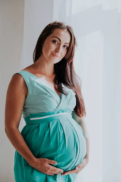 Zwangere vrouw in een witte kamer staat bij het raam — Stockfoto