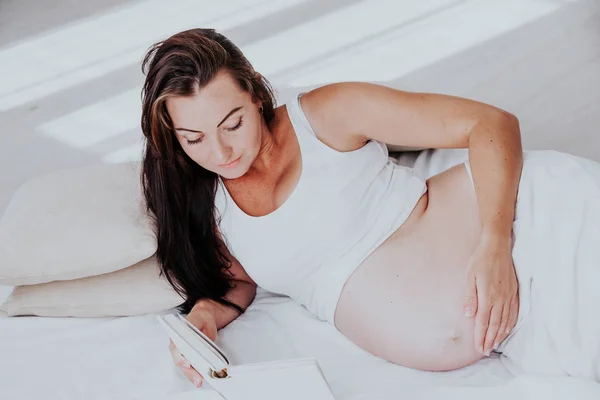 Una mujer embarazada yace en una cama y lee un libro —  Fotos de Stock