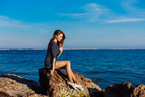 The girl in the striped dress sits on the rocks by the sea — Stock Photo, Image