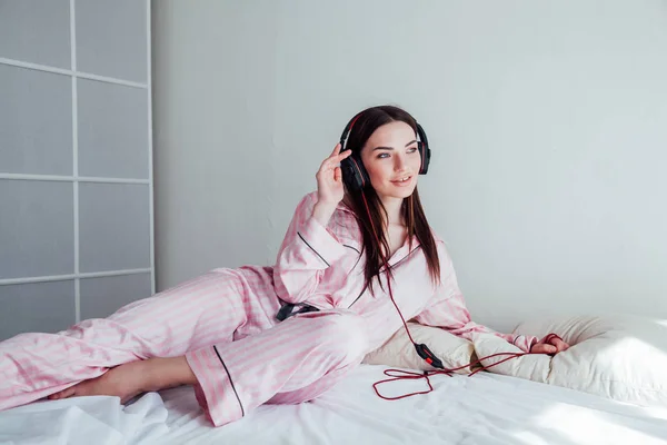 ピンクのパジャマを着て、ベッドの上にヘッドフォンで音楽を聴くブルネット — ストック写真