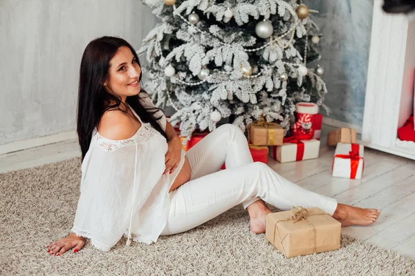 Ciężarna kobieta siedząca przy choince otwiera prezenty świąteczne — Zdjęcie stockowe