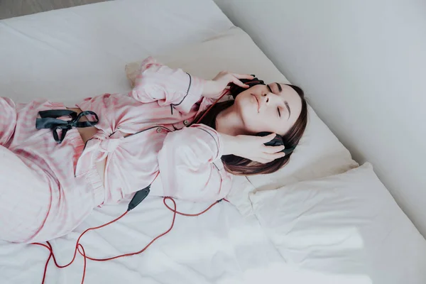 Vrouw in roze pyjama's, luisteren naar muziek in hoofdtelefoons dansen op het bed — Stockfoto