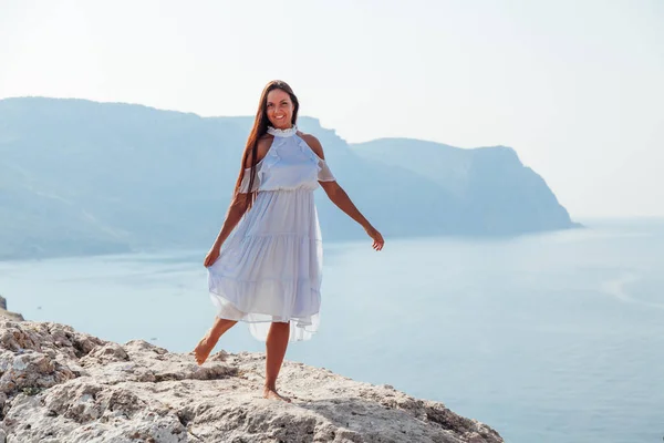Beautiful tanned woman in blue dress on top of a cliff by the sea — Stock Photo, Image