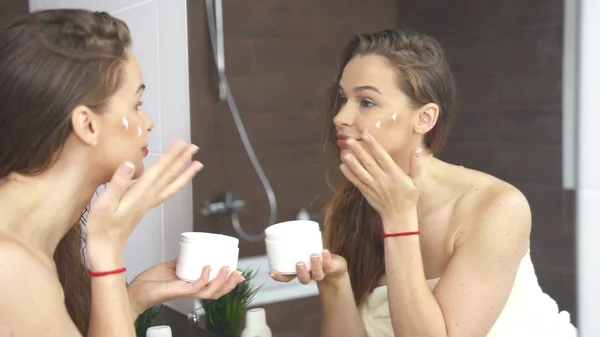 Woman applying cream to skin while face makeup at mirror in bathroom.