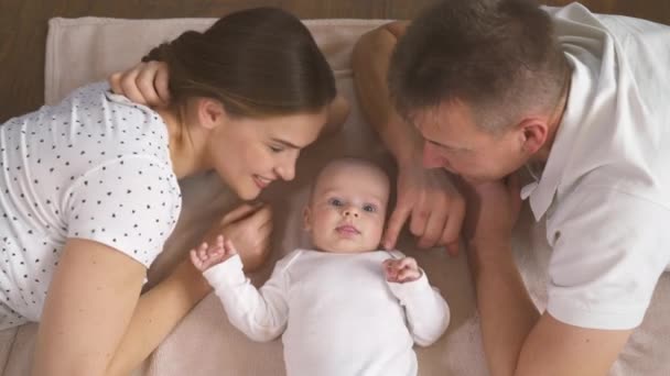 Glückliche Familie. Mama Papa und drei Monate altes Baby. Familie liegt im Bett und blickt lächelnd in die Kamera. — Stockvideo