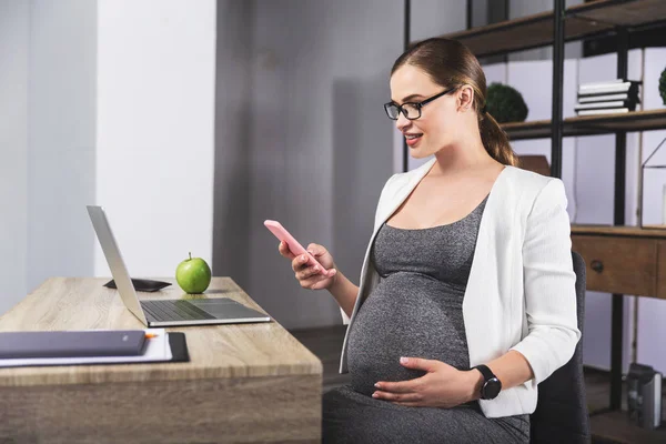Mujer embarazada joven mirando por teléfono mientras trabaja en la oficina — Foto de Stock