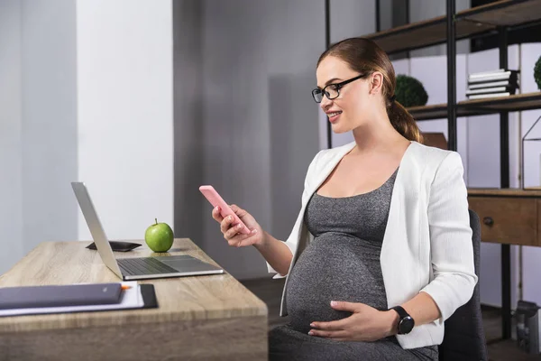 Mujer embarazada joven mirando por teléfono mientras trabaja en la oficina — Foto de Stock