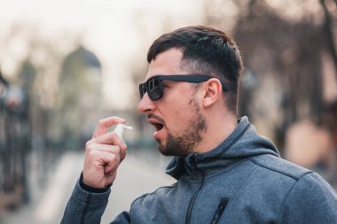 Yakışıklı sakallı bir adam boğazı için öksürük spreyi kullanır ve ağzını kocaman açar. Sokağın arka planında güneş gözlüklü bir portre. Reklam, koronavirüs, hastalık, enfeksiyon, karantina, covid-19