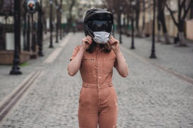 Tulum giymiş ince bir kız. Tıbbi maskeli bir kadın motosiklet kaskı takıyor. Yüzü olmayan fotoğraf. Caddenin ve yolun arka planına karşı. Koronavirüs, hastalık, enfeksiyon, karantina, covid-1