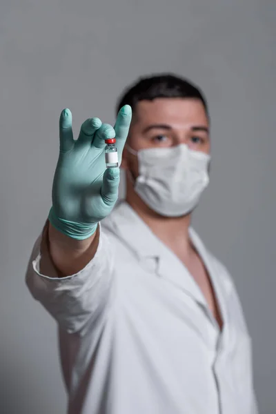 A  beautiful doctor in a White medical gown is holding in his hand with glove a jar of pills or injection with a medical mask on his face