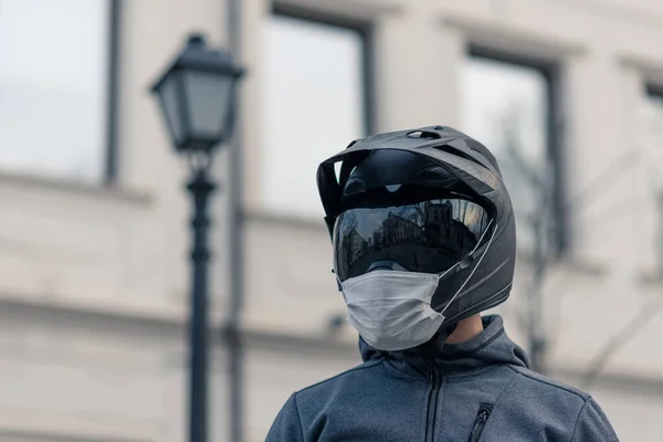 Kasklı bir adamın portresi. Motosiklet kaskında beyaz bir tıbbi maske. Sokakta çekilmiş bir fotoğraf, arka plandaki evlere karşı. Koronavirüs, hastalık, enfeksiyon, karantina, covid-19