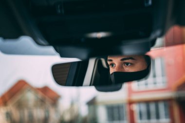 Siyah tıbbi maskeli bir adam. Yakışıklı esmer, araba kullanıyor. Araba aynasında fotoğraf var. Hırsız, hırsız, koronavirüs, hastalık, enfeksiyon, karantina, tıbbi maske, covid-19