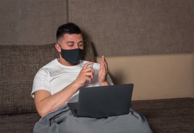 Tıbbi maskeli beyaz bir adam, Coronavirus veya Covid-19 enfeksiyonu sırasında evden bilgisayar kullanarak karantina servisindeki bir hastayla çalışır. Bacaklarını yorganın altına koy. ev yalıtımı