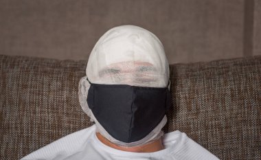 Sakallı bir adamın sargılı kafası. Kafa sargı bezine sarılmış. Görünürde kimse yok. Yüzünde siyah bir maske var. Evde ilk yardım. Coronavirus, covid 19, karantina, mumya.