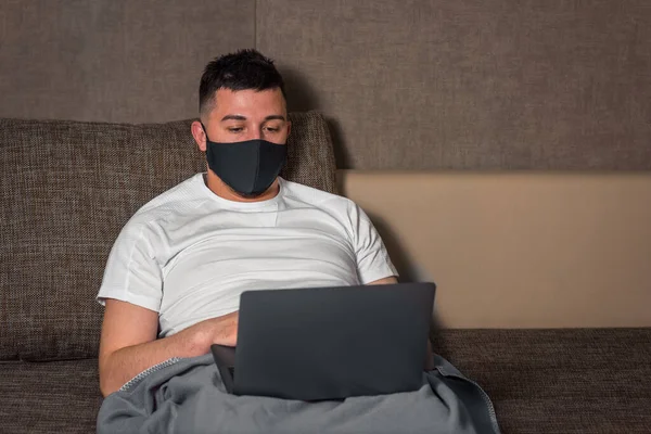 Tıbbi maskeli beyaz bir adam, Coronavirus veya Covid-19 enfeksiyonu sırasında evden bilgisayar kullanarak karantina servisindeki bir hastayla çalışır. Bacaklarını yorganın altına koy. ev yalıtımı