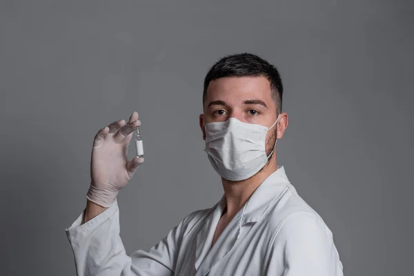 白い医療用ガウンを着た男顔と手術用手袋のマスク白色または灰色の背景にワクチンのガラス瓶を見て — ストック写真