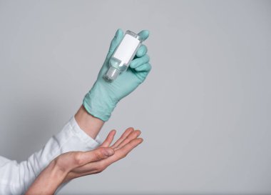 Bulaşıcı virüslere, bakterilere ve mikroplara karşı korunmak için dezenfektan ya da antiseptik jel uygulayan mavi bir tıbbi eldivenin içindeki bir el gri arka planda eldivensiz bir ele dökülür.