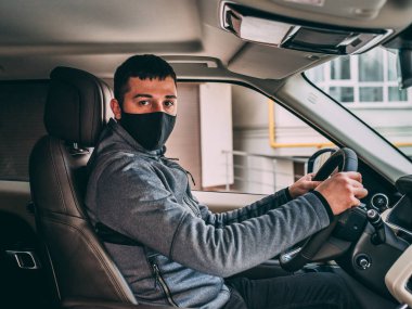 Siyah tıbbi maskeli bir adam. Yakışıklı esmer, pahalı bir arabanın direksiyonunda oturuyor. Hırsız, hırsız, koronavirüs, hastalık, enfeksiyon, karantina, tıbbi maske, covid-19