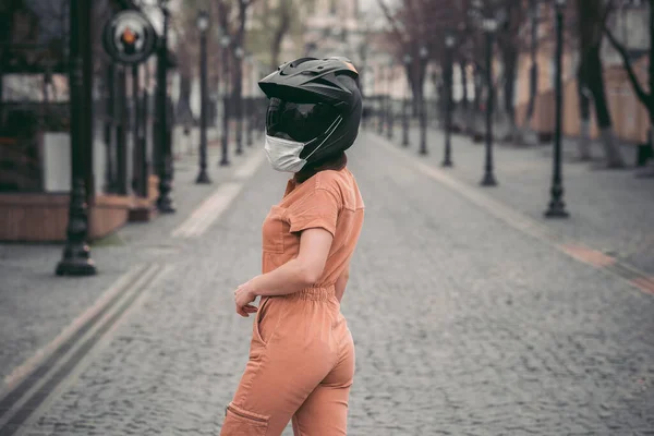 slender girl in overalls. A woman is standing in a motorcycle helmet with a medical mask. Photo without a face. Against the background of the street and road. coronavirus, disease, infection, quarantine, covid-1
