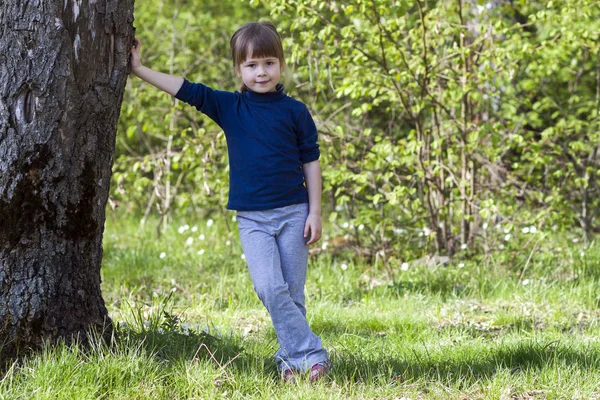 Sevimli gülümseyen küçük kız yeşil çimenlerin üzerinde büyük ağacın yanında ayakta — Stok fotoğraf