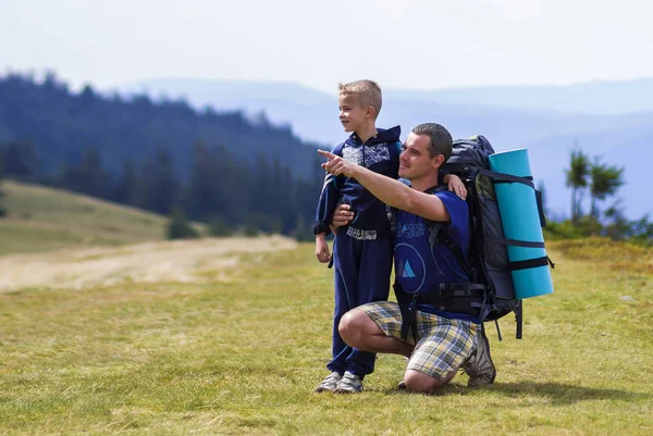 Vater und Sohn wandern mit Rucksäcken gemeinsam in den sommergrünen Bergen. Vater und Kind stehen und genießen die Aussicht auf die Berge. Aktiver Lebensstil, Familienbeziehungen, Wochenendaktivitätskonzept. — Stockfoto