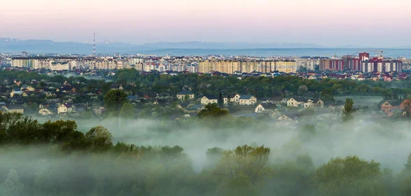 Hermoso panorama de la mañana vista aérea de la ciudad de Ivano-Frankivsk, Ucrania. Escena de suburbios tranquilos entre árboles verdes bajo densa niebla y edificios altos en lejanos Cárpatos montañas de fondo . — Foto de Stock