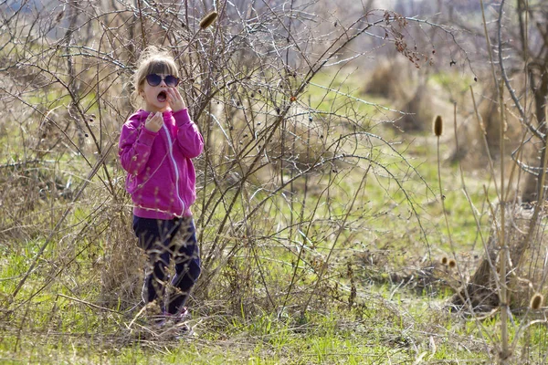 Porträtt av söt liten förvirrad blond flicka i rosa fritidskläder och mörka solglasögon står ensam borttappad bland torra taggiga buskar och kallar på hjälp. Rädslor och faror i barn fantasi. — Stockfoto