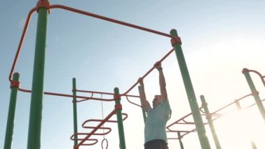 Aktif sporlarda güçlü vücut eğitimi almış genç sporcu açık havada yatay fitness barına yanaşıyor..