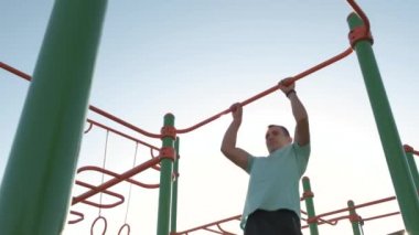 Aktif sporlarda güçlü vücut eğitimi almış genç sporcu açık havada yatay fitness barına yanaşıyor..