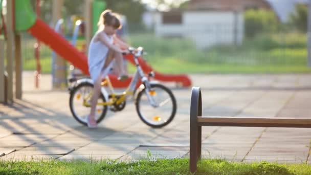 かわいい女の子がベンチに座って夏に学校の庭で自転車に乗って — ストック動画