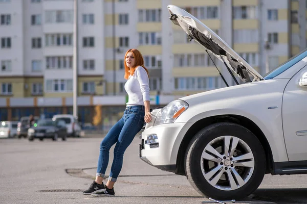 Young stressed woman driver standing near broken car with open h