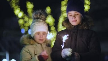 İki tatlı genç çocuk, sıcak kış kıyafetleri içinde kız ve erkek, açık havada karanlık bir gecede havai fişek yakıyorlar. Yeni yıl ve Noel kutlaması konsepti.