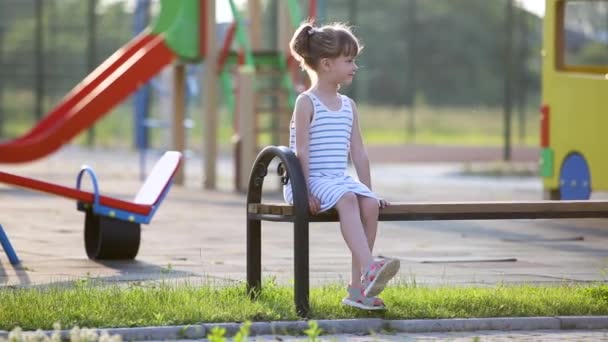 可爱的小女孩坐在幼儿园操场上的长椅上等妈妈 — 图库视频影像