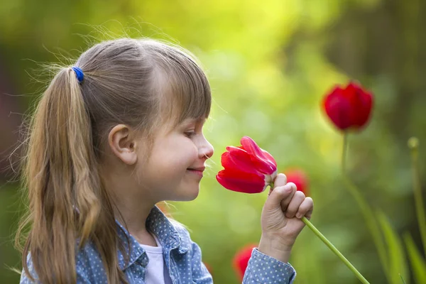 Perfil da menina bonita e sorridente criança com olhos grisalhos e cabelos longos cheirando flor de tulipa vermelha brilhante no verão ensolarado borrado fundo bokeh verde . — Fotografia de Stock