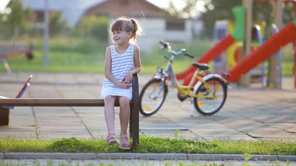 かわいい女の子がベンチに座って夏に学校の庭で自転車に乗って — ストック動画