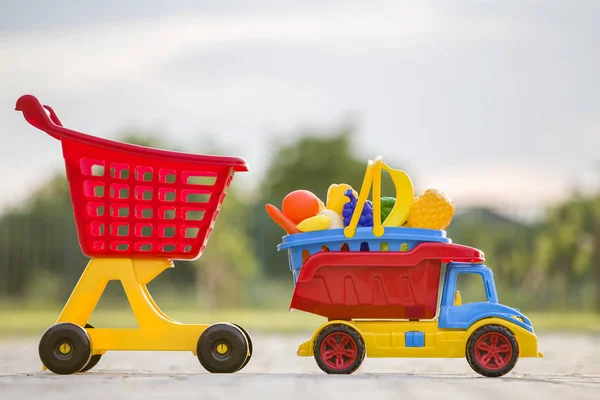Jasne plastikowe kolorowe zabawki dla dzieci na świeżym powietrzu w słoneczny letni dzień. Samochód ciężarowy niosący koszyk z zabawkami owocowo-warzywnymi i wózkiem sklepowym. — Zdjęcie stockowe