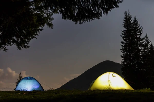 Two brightly lit tourist tents on green grassy forest clearing on dark mountain and clear blue starry sky copy space background. Tourism, night camping in summer mountains, beauty of nature concept.