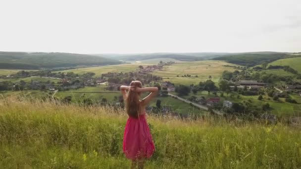 田舎の丘の環境で背の高い緑の草と夏のフィールドで歩く赤いドレスで長い髪を持つ龍の女性 — ストック動画