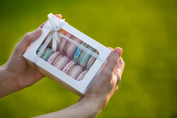 Manos femeninas sosteniendo caja de regalo de cartón con coloridas galletas macaron hechas a mano de color azul rosado sobre fondo de copia borrosa verde . — Foto de Stock