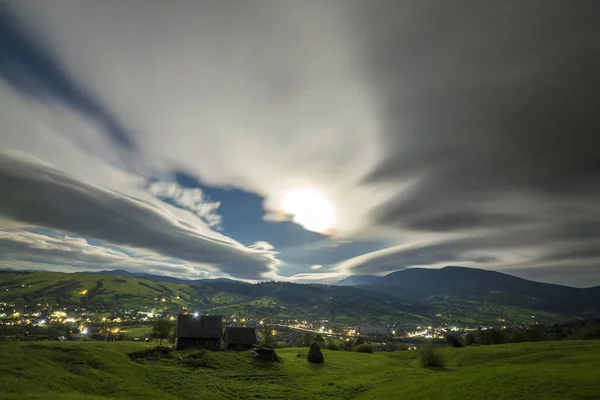 Noche de verano panorama de montaña. Viejas cabañas de pastor envejecidas de madera en el claro verde en el fondo nublado del cielo de la noche, camino brillante y luces de vivienda en el fondo del valle . — Foto de Stock