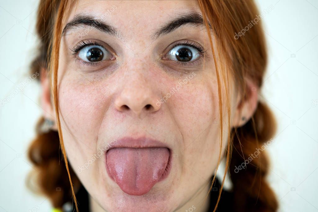 Closeup portrait of a funny redhead teenage girl with childish hairstyle sticking out her tongue isolated on white backround.