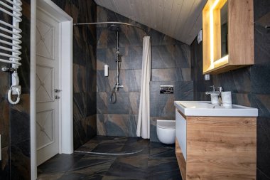 Modern tarz banyo, siyah fayanslı duvarlar, perde duş yeri ve leğen ve büyük ışıklı aynalı ahşap mobilyalar..