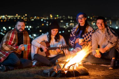 Bir grup insan gece dışarıda şenlik ateşinin yanında oturup gitar çalarak, şarkı söyleyerek ve birlikte mutlu bir şekilde konuşarak eğleniyor..